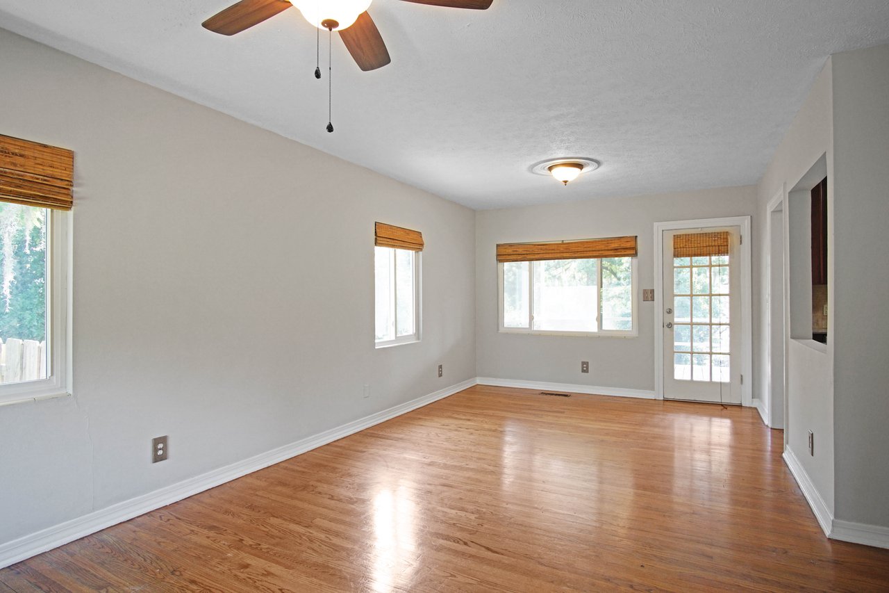 Bright, empty room with hardwood floors, white walls, and two windows with wooden blinds. A ceiling fan and a glass door add to its airy feel.