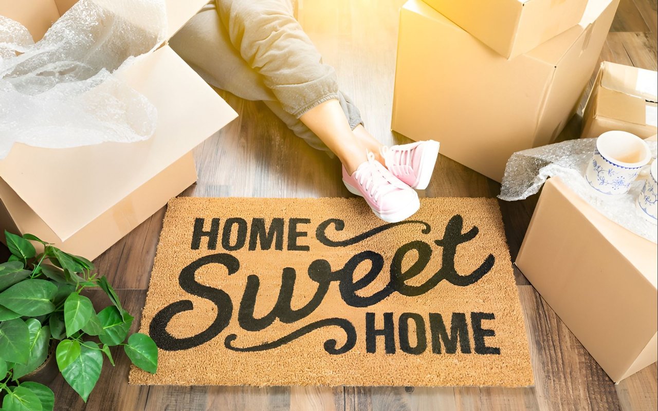 A person among moving boxes rests pink sneakers on a “HOME Sweet HOME” doormat.