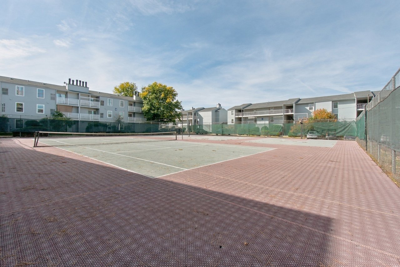 Outdoor tennis court beside multi-story apartment buildings, with a basketball hoop and fenced perimeter.