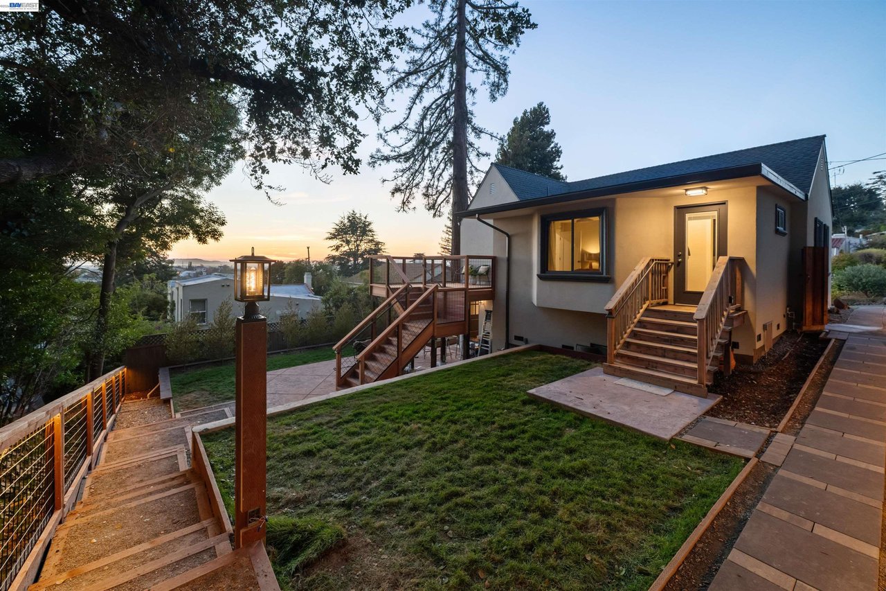 Gray house above a landscaped backyard, with wooden stairs and a raised deck at dusk.