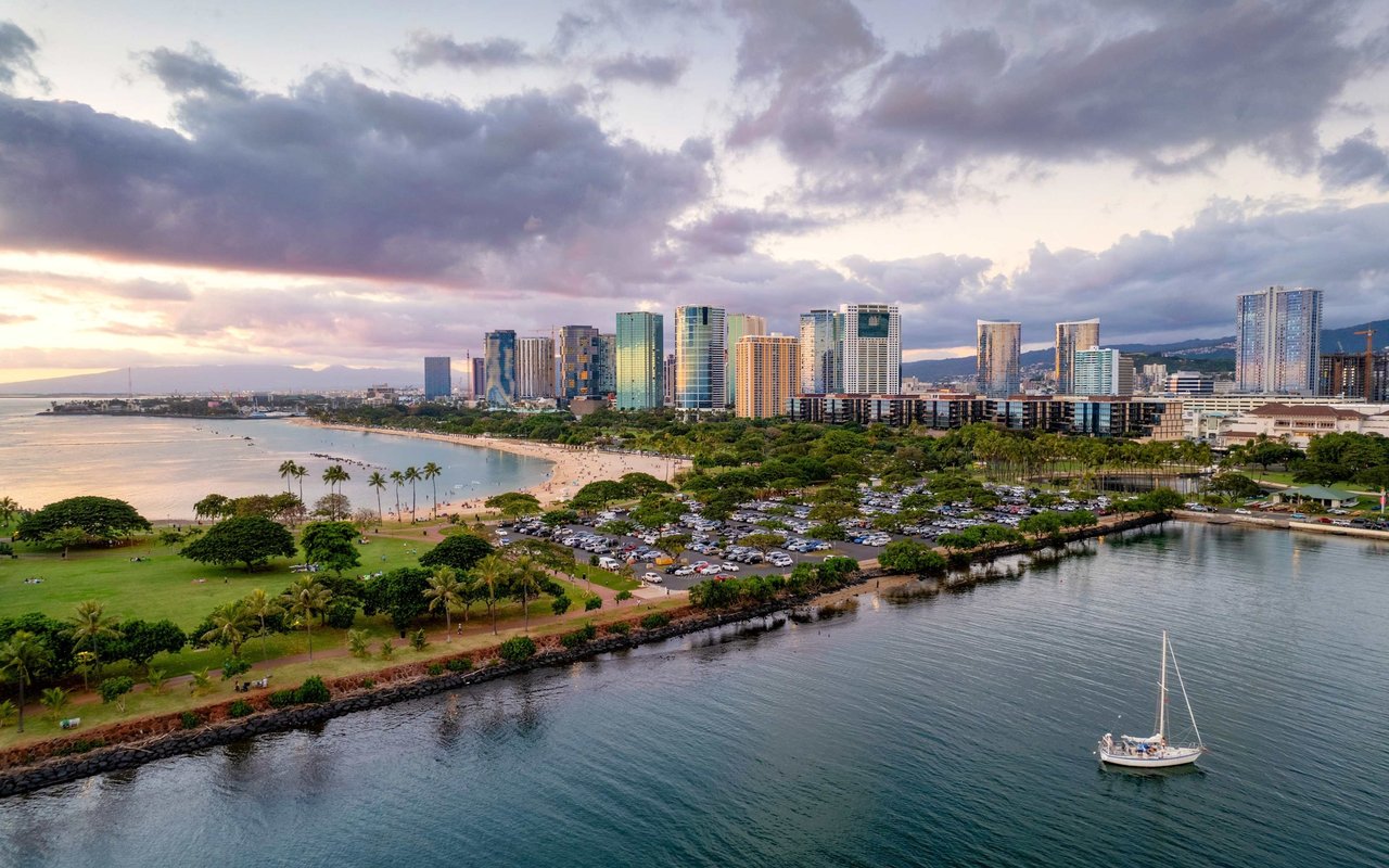 Kaka'ako waterfront skyline at sunset, with beach, parkland, and a sailboat in the harbor.
