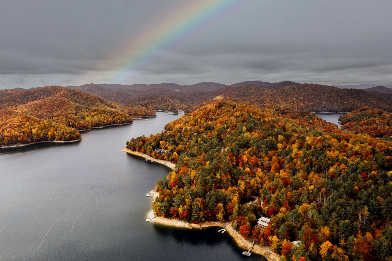 Alt=Aerial view of a lake surrounded by forested hills with autumn foliage. A rainbow arches over the cloudy sky, and several docks, are visible along the shoreline.