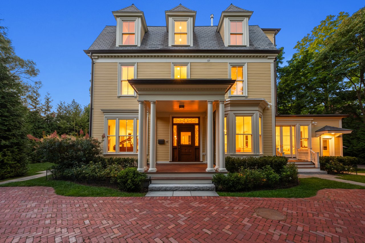 Victorian house with three dormers, a columned portico, glowing windows, and a brick driveway at dusk.