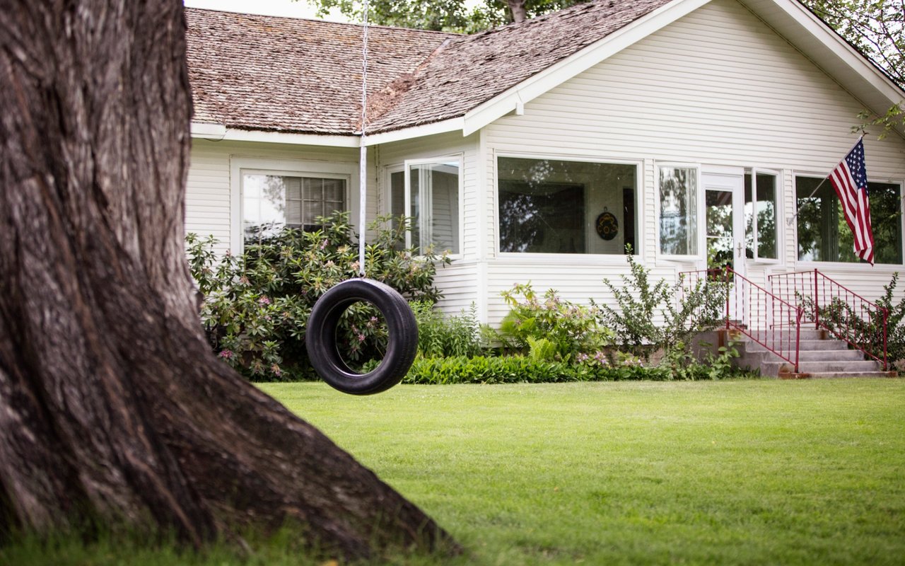 White-sided house with red step railings and an American flag, seen beyond a lawn and tire swing.