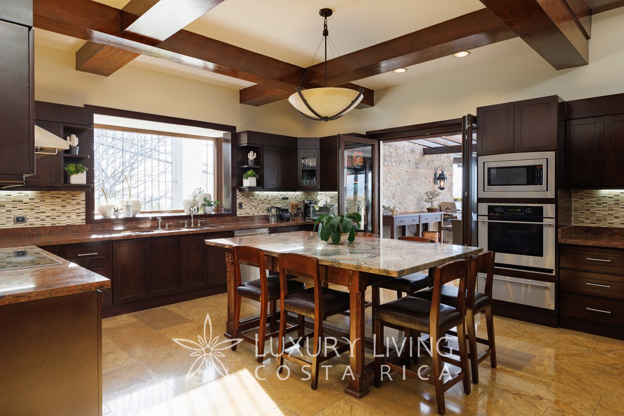 Luxury kitchen with dark wood cabinetry, stone countertops, and a large dining island beneath exposed beams.