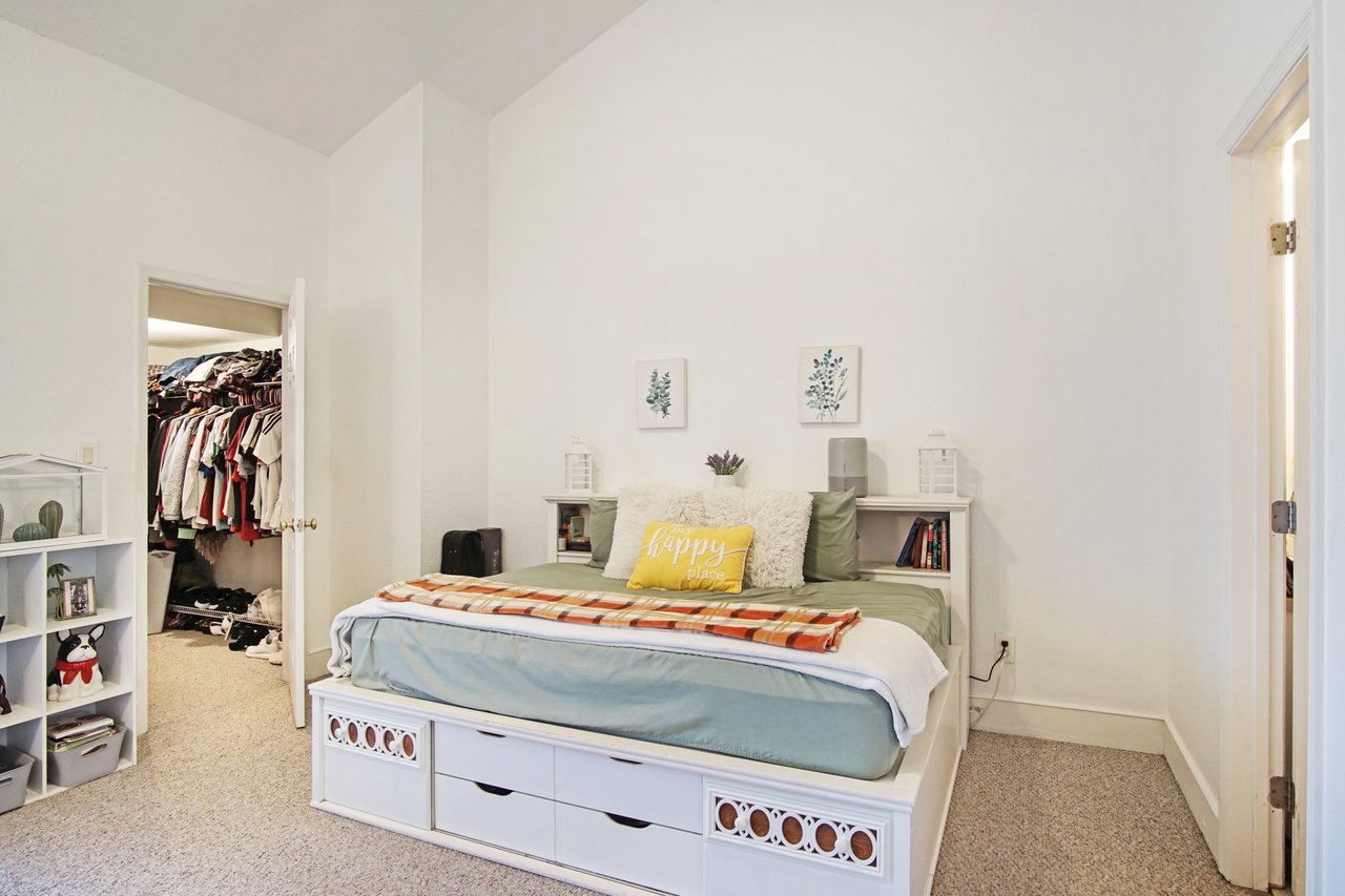 Spacious bedroom with a bed featuring green bedding and a yellow pillow reading "happy place." An open closet displays organized clothes. Cozy and inviting.