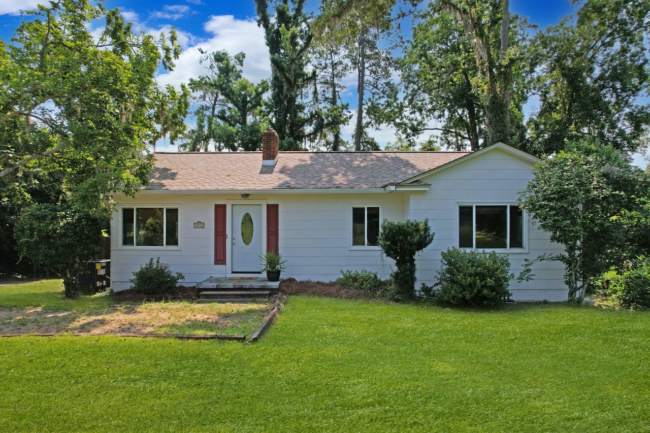 Single-story white house with a red door, surrounded by lush green grass and trees. Bright blue sky with a few clouds, conveying a peaceful, sunny day.