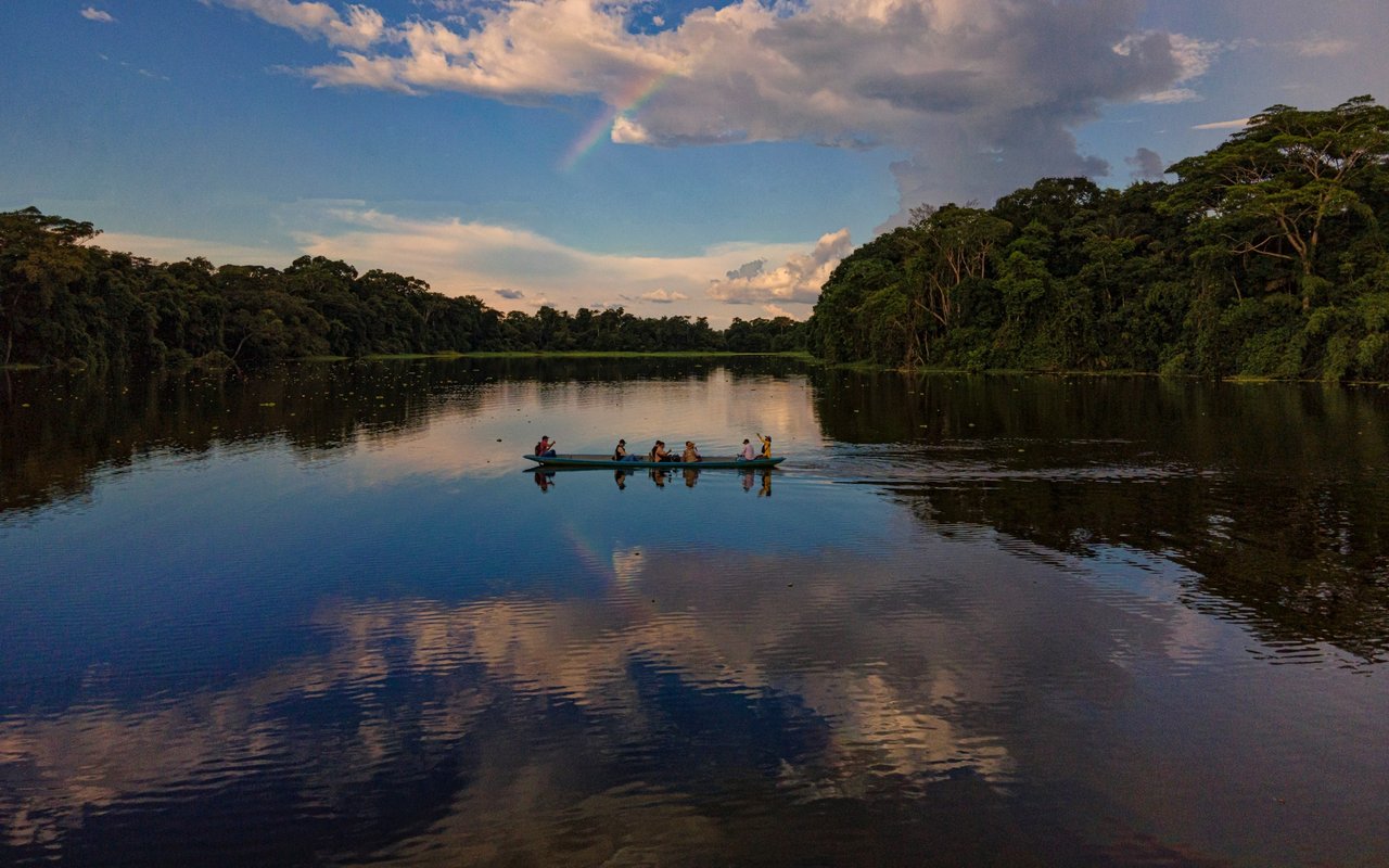 Forest-lined river with passengers in a canoe beneath a rainbow.