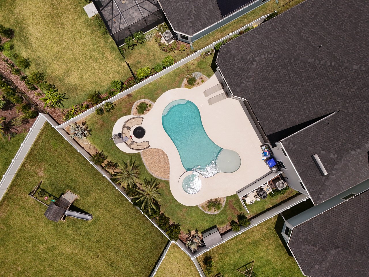 Aerial view of a backyard pool with a large patio, outdoor seating, and fenced lawn.