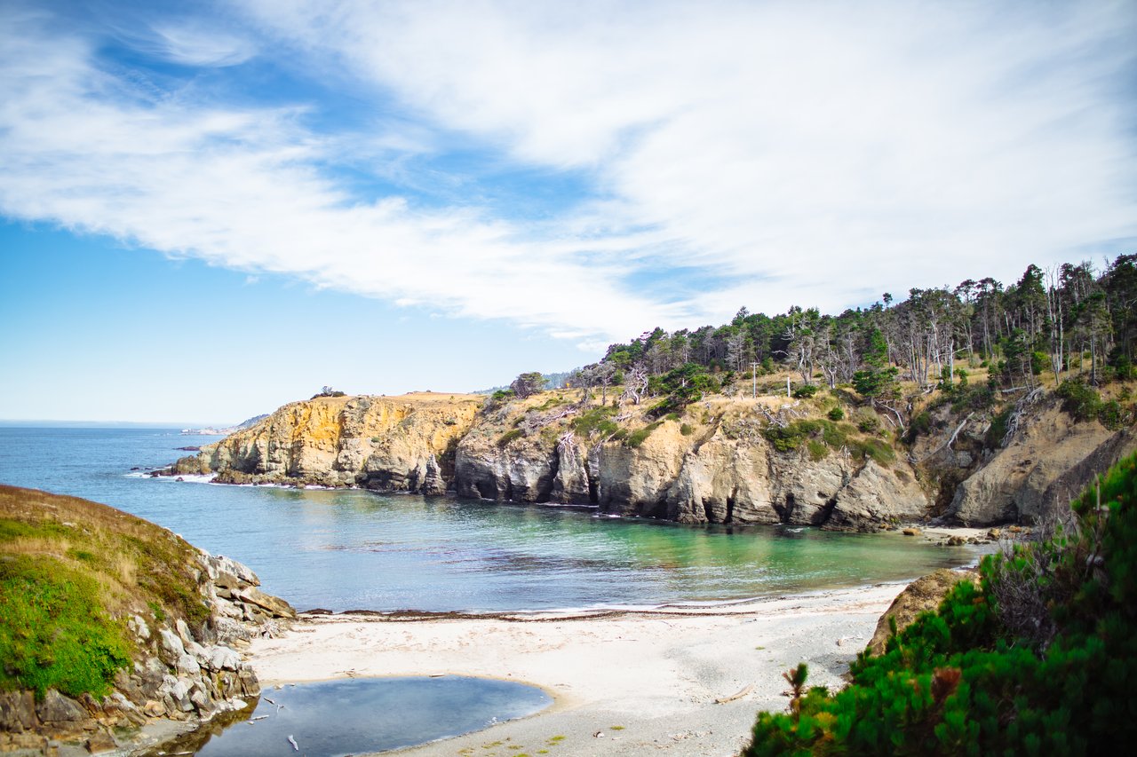 A sandy cove beneath rugged, tree-topped cliffs along the ocean.
