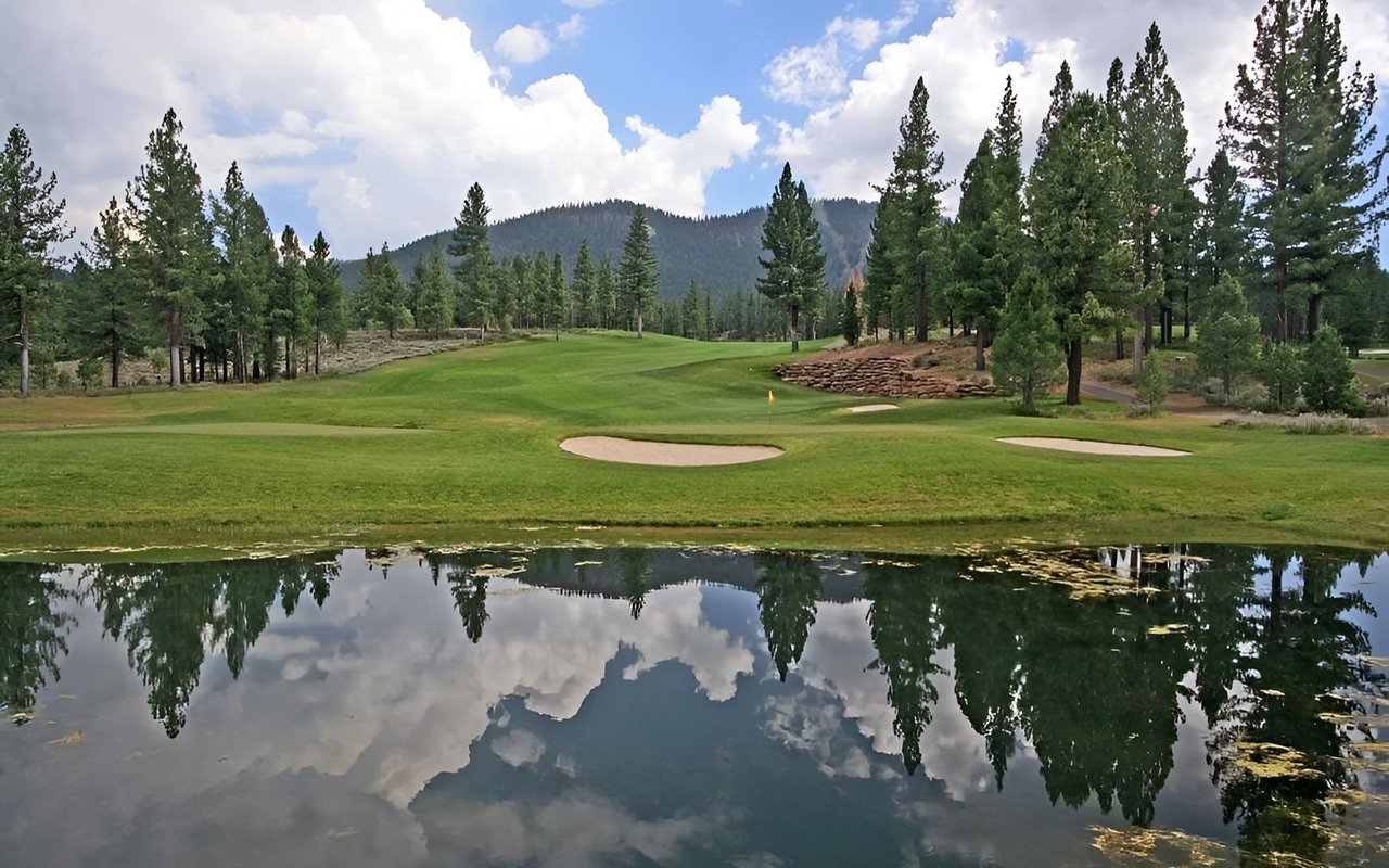 Golf course beside a reflective pond, with pine trees and mountains beyond.