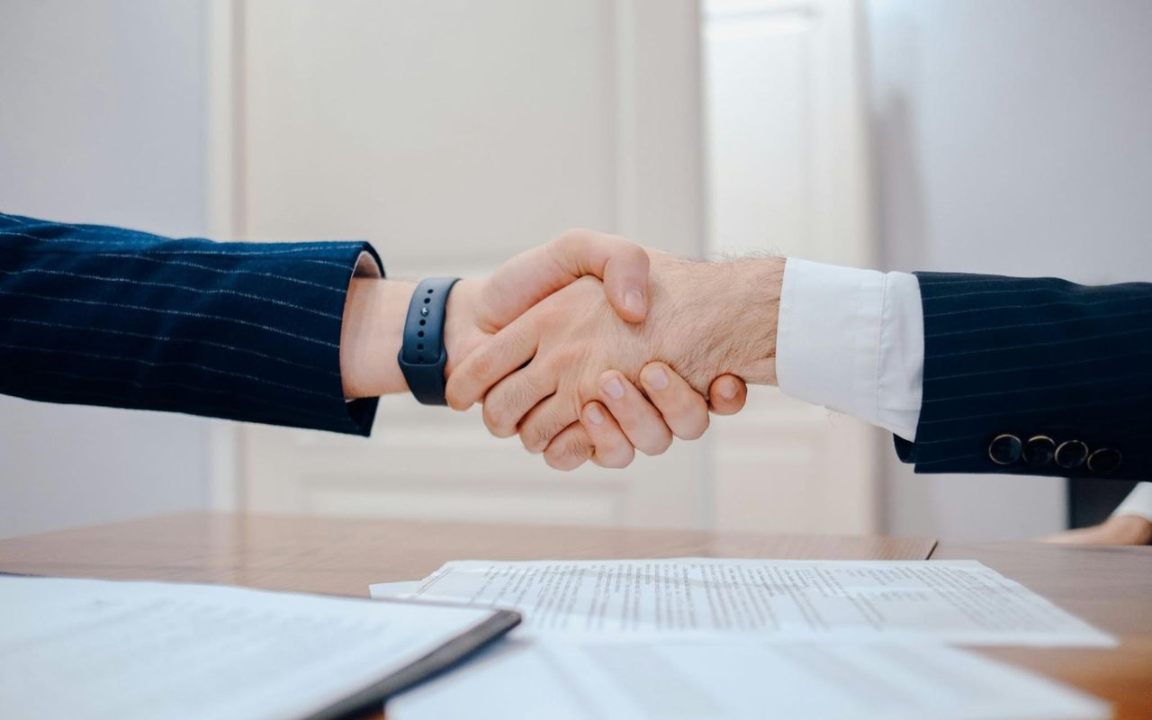 Close-up of a business handshake between two individuals in pinstripe suits over paper contracts on a wooden desk.