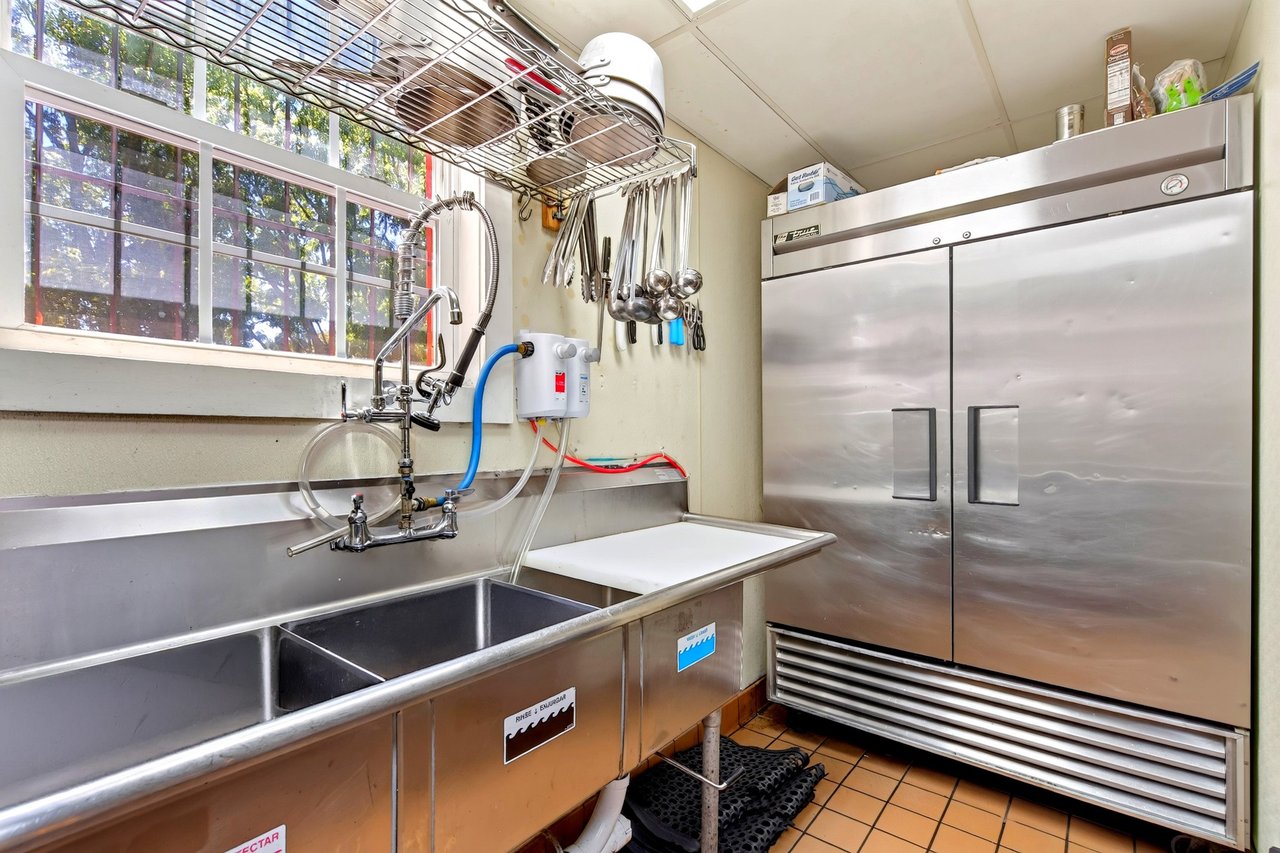 Restaurant kitchen with a commercial double sink, hanging utensils, and a large stainless-steel refrigerator.
