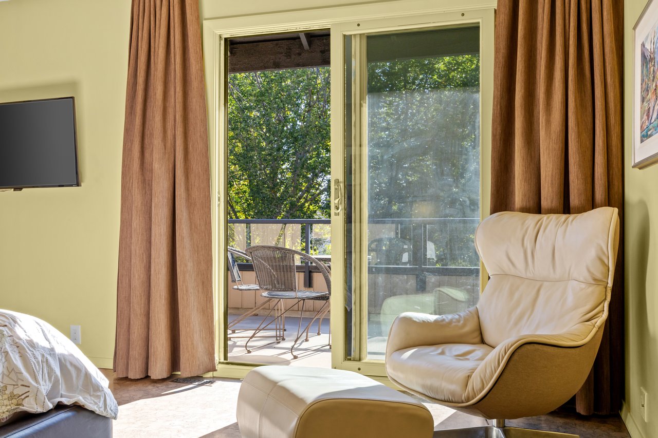 Bedroom sitting area with a cream leather chair beside balcony doors and leafy trees outside.