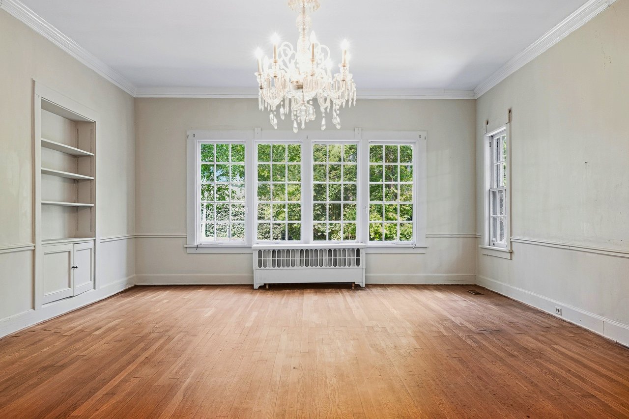 Empty room with hardwood floors, built-in shelves, a crystal chandelier, and large windows overlooking trees.