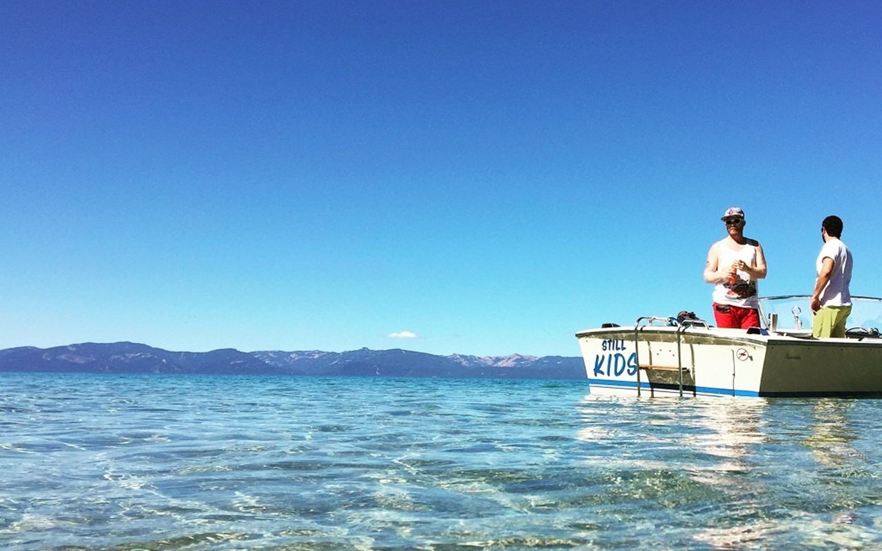 Two men aboard a small boat on clear Lake Tahoe water, with mountains in the distance.