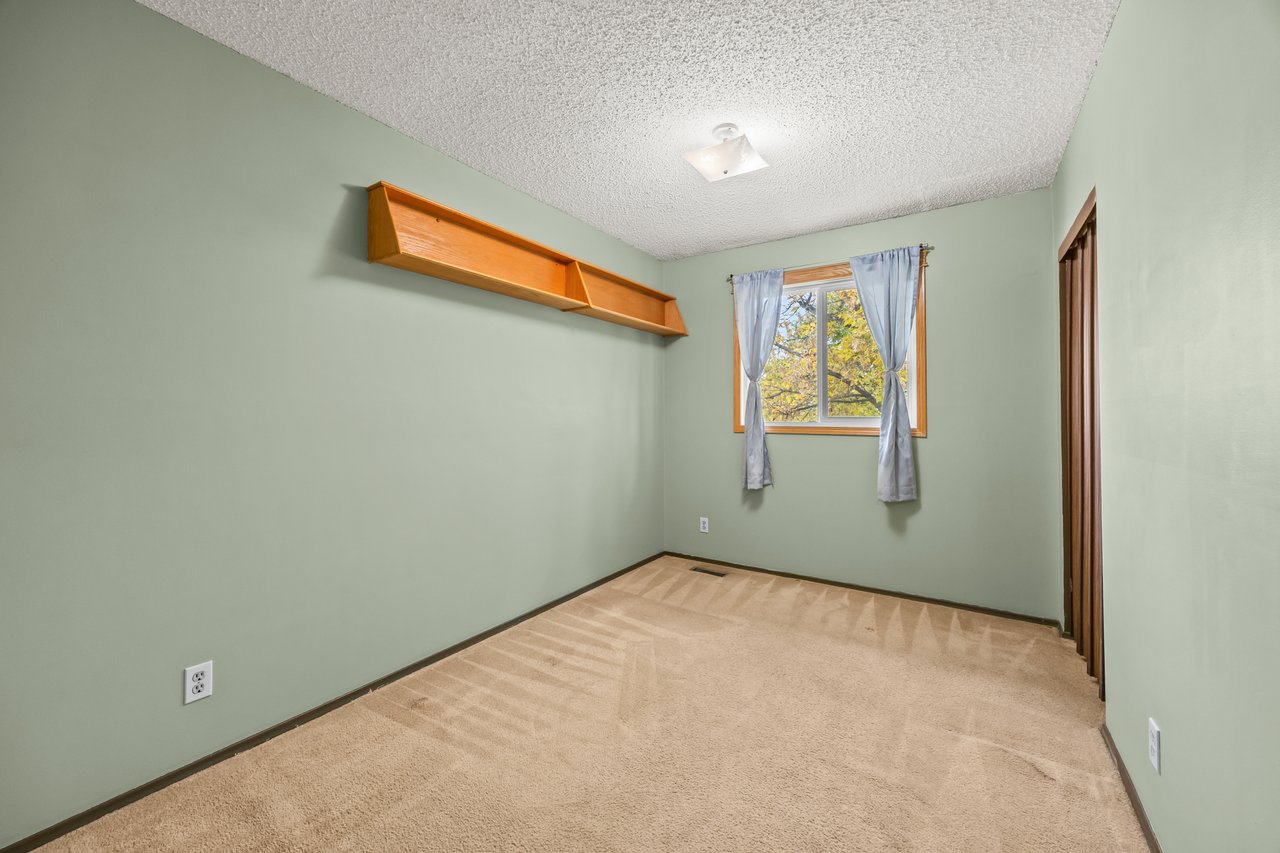 Empty bedroom with pale green walls, beige carpet, a wooden wall shelf, and a curtained window.