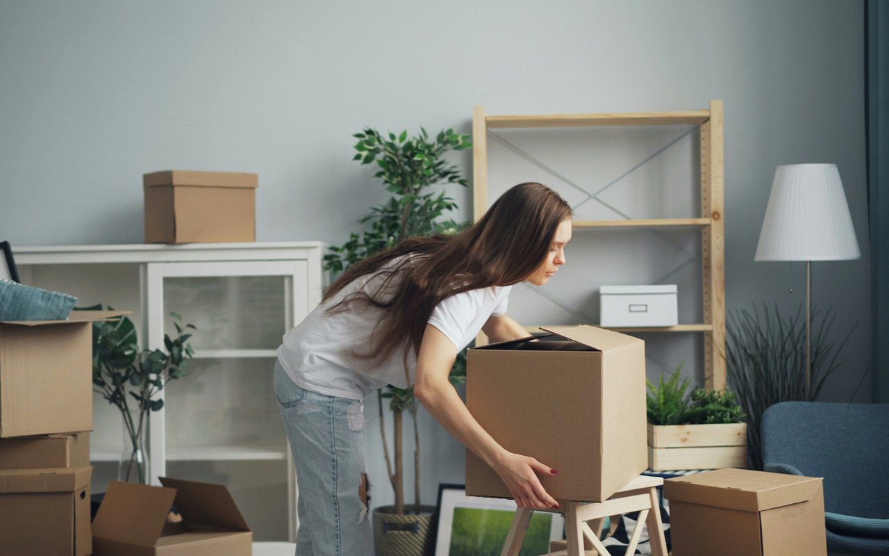 Woman packs a large cardboard box in a furnished room surrounded by moving boxes.