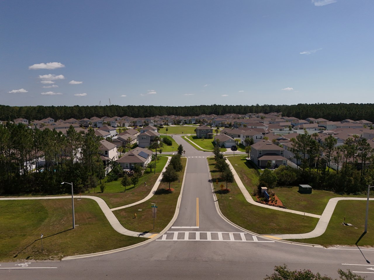 Aerial view of a residential neighborhood with a central road, sidewalks, and a wooded tree line.