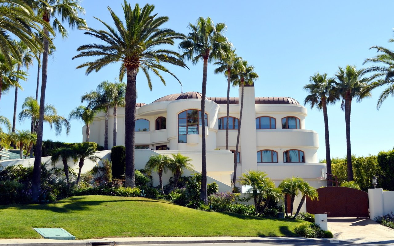 White rounded residence framed by tall palms, with a green lawn and gated driveway.