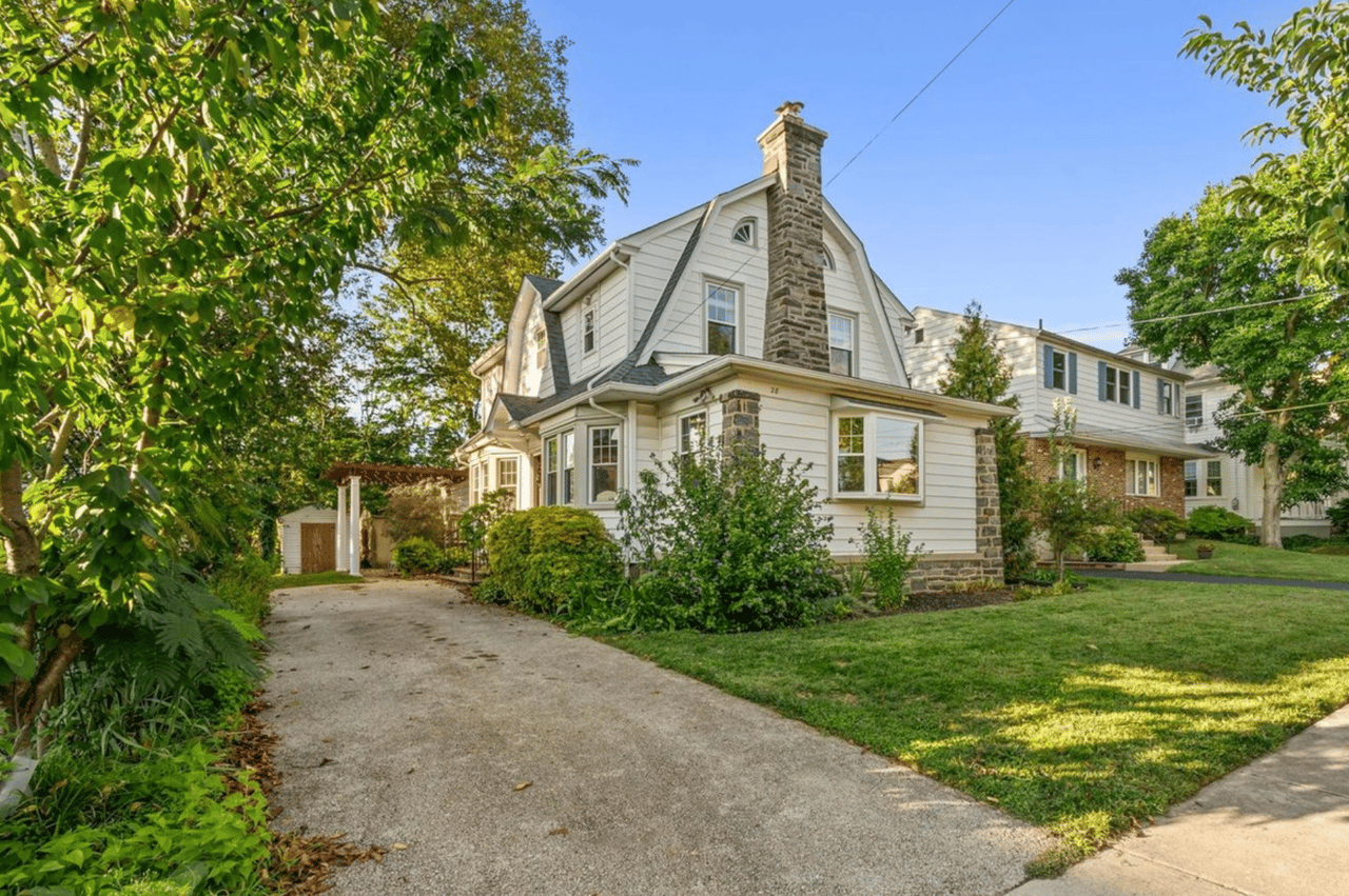 White clapboard house with a tall stone chimney, surrounded by mature trees and gardens.