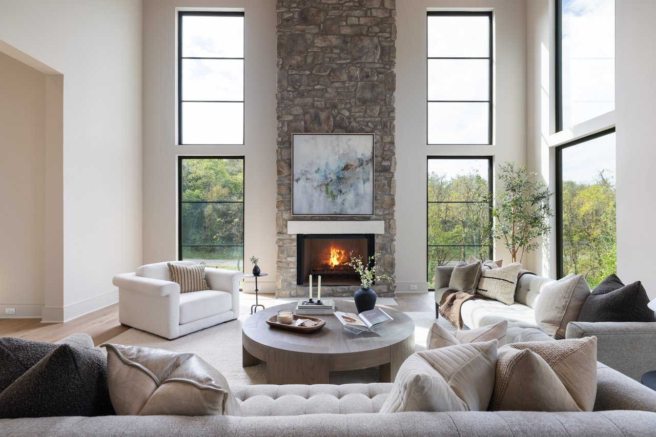 Living room with a tall stone fireplace, high windows overlooking trees, and neutral upholstered sofas.