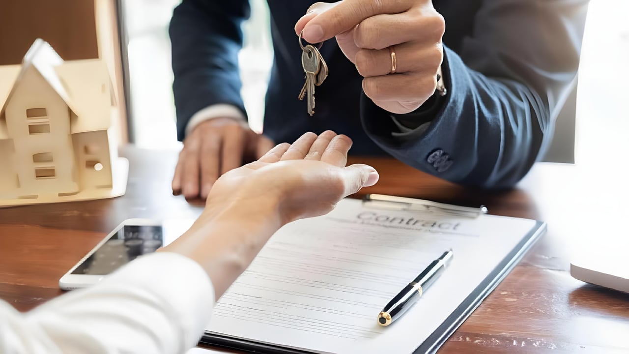 Hands exchange house keys over a real estate contract beside a model home.