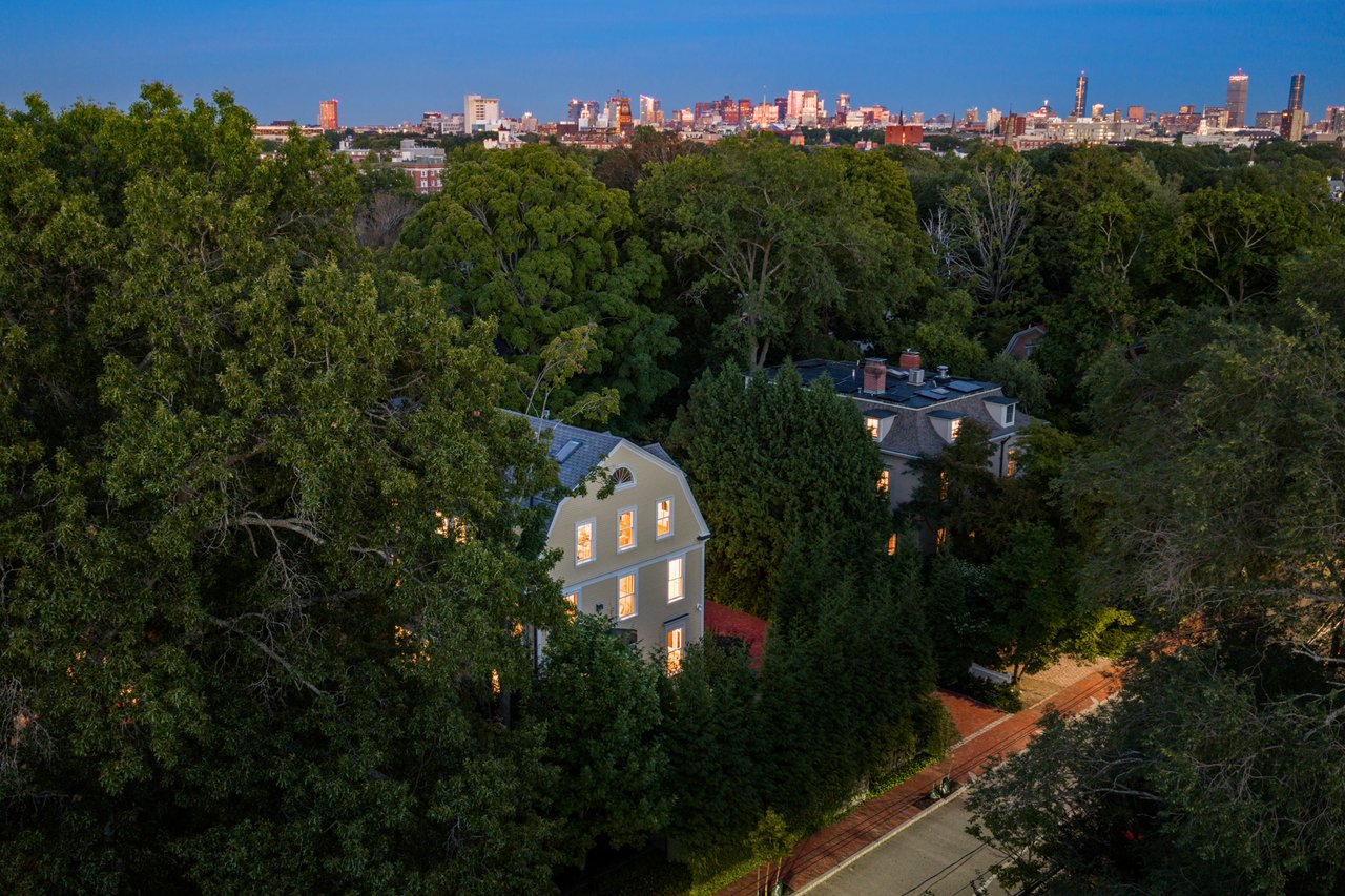 Aerial view of a tree-surrounded Victorian home with the illuminated city skyline beyond.