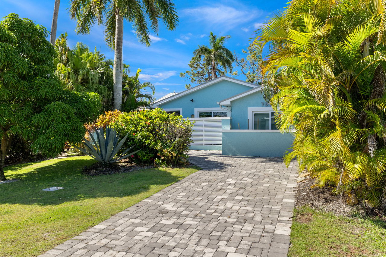 Blue single-story home with a paver driveway, low wall, and lush palms.