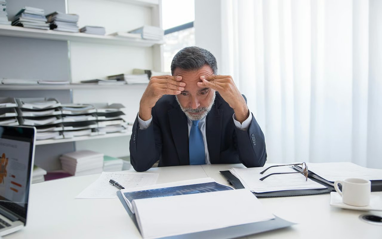 Stressed man in a suit studies paperwork at a cluttered office desk.