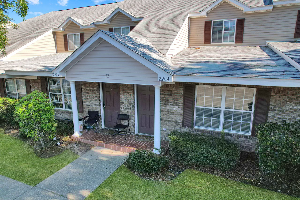 A brick townhouse with a gabled roof features two doors numbered 2203 and 2204. Two chairs sit on the porch, with tidy green shrubs and a path in the foreground. Bright and sunny day.