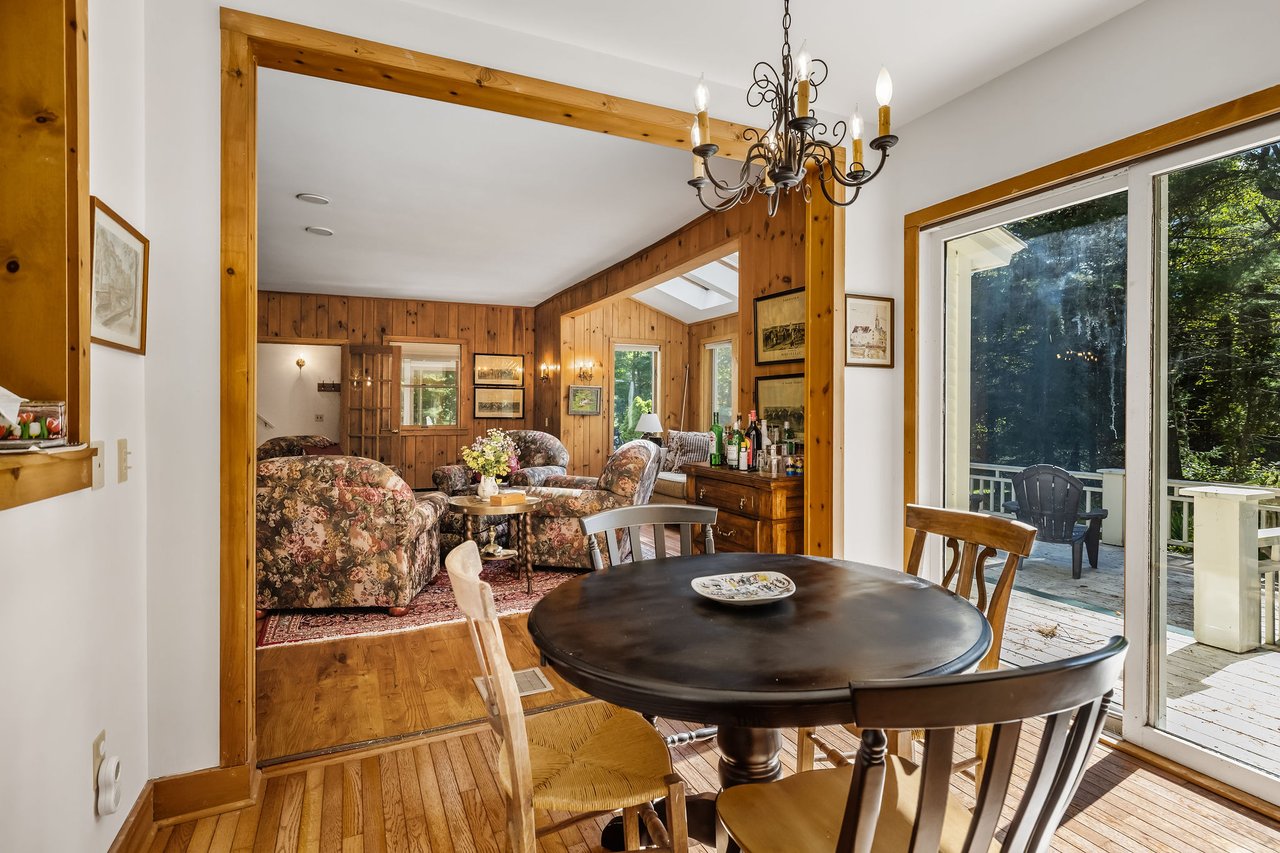 Dining room with a round dark wood table, adjoining wood-paneled living room, and glass doors to a wooded deck.