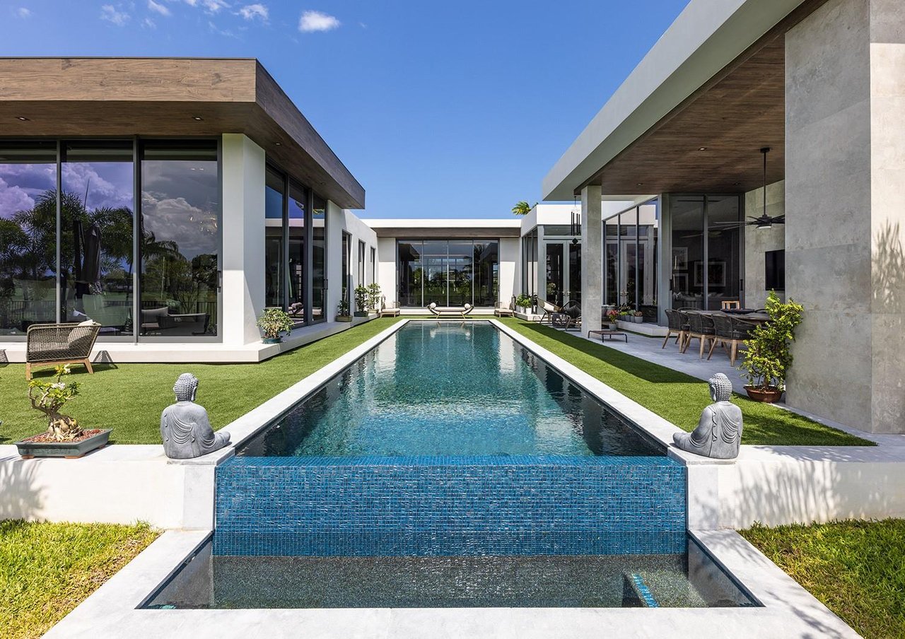 Modern home surrounding a long pool, with floor-to-ceiling glass, wood-paneled eaves, and outdoor seating.