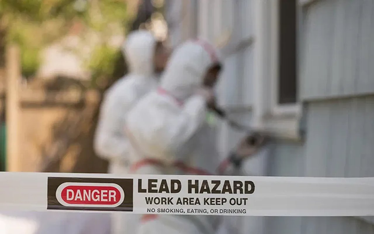 Lead hazard warning tape in front of a worker in protective gear at a house window.