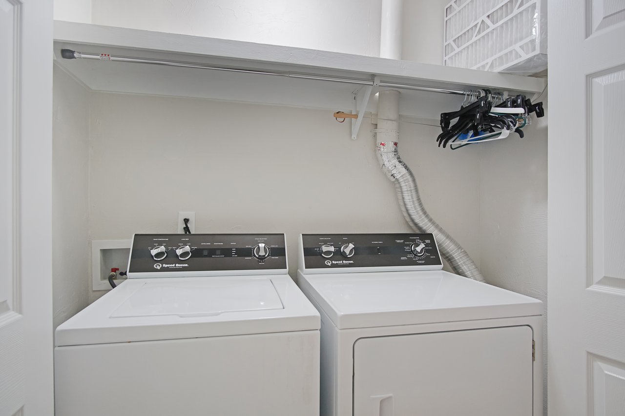 Laundry room with a white washer and dryer, a hanging rod above with clothes hangers, and a metal ventilation hose. Bright, organized space.