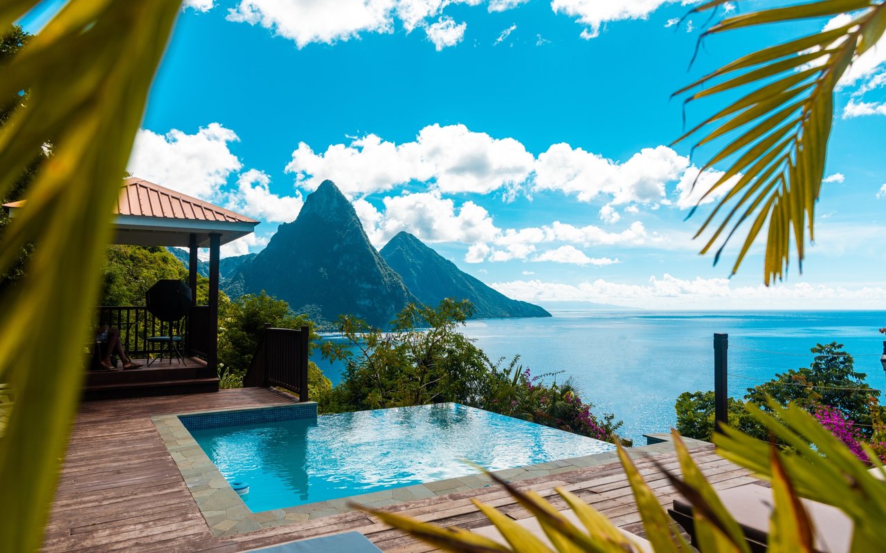 Pool and wooden deck overlooking the ocean and steep green mountains, framed by palm fronds.