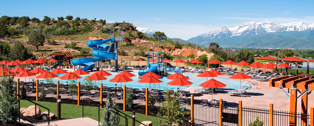 Outdoor pool with blue waterslides and red umbrellas beneath snow-capped mountains.