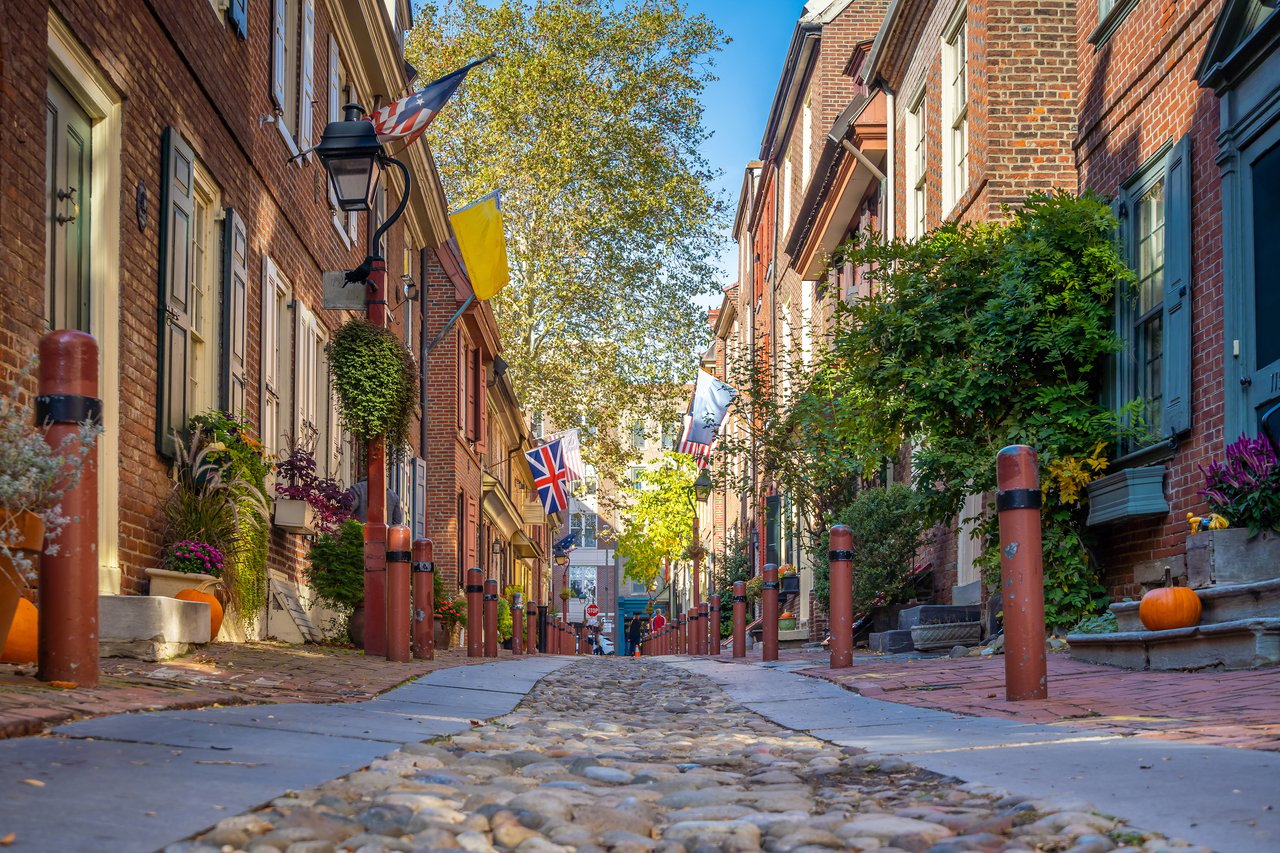 Cobblestone alley between brick row houses, decorated with flags, potted plants, and pumpkins.