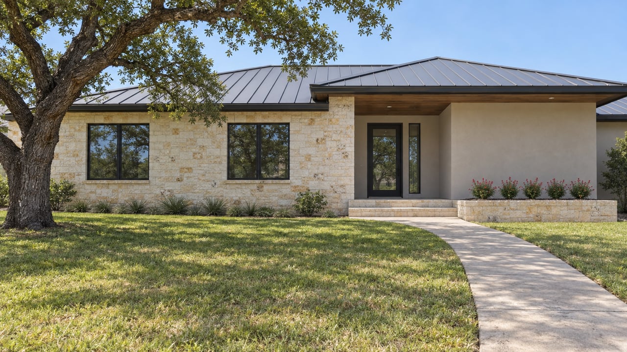 Sunlit limestone and stucco home with a charcoal metal roof, deep front lawn and spreading live oak.