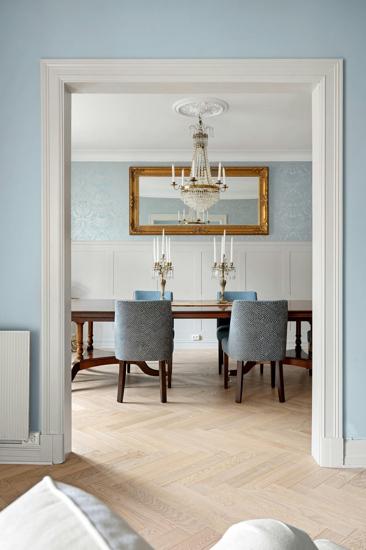 Dining room with crystal chandeliers, a gold-framed mirror, and blue patterned chairs.
