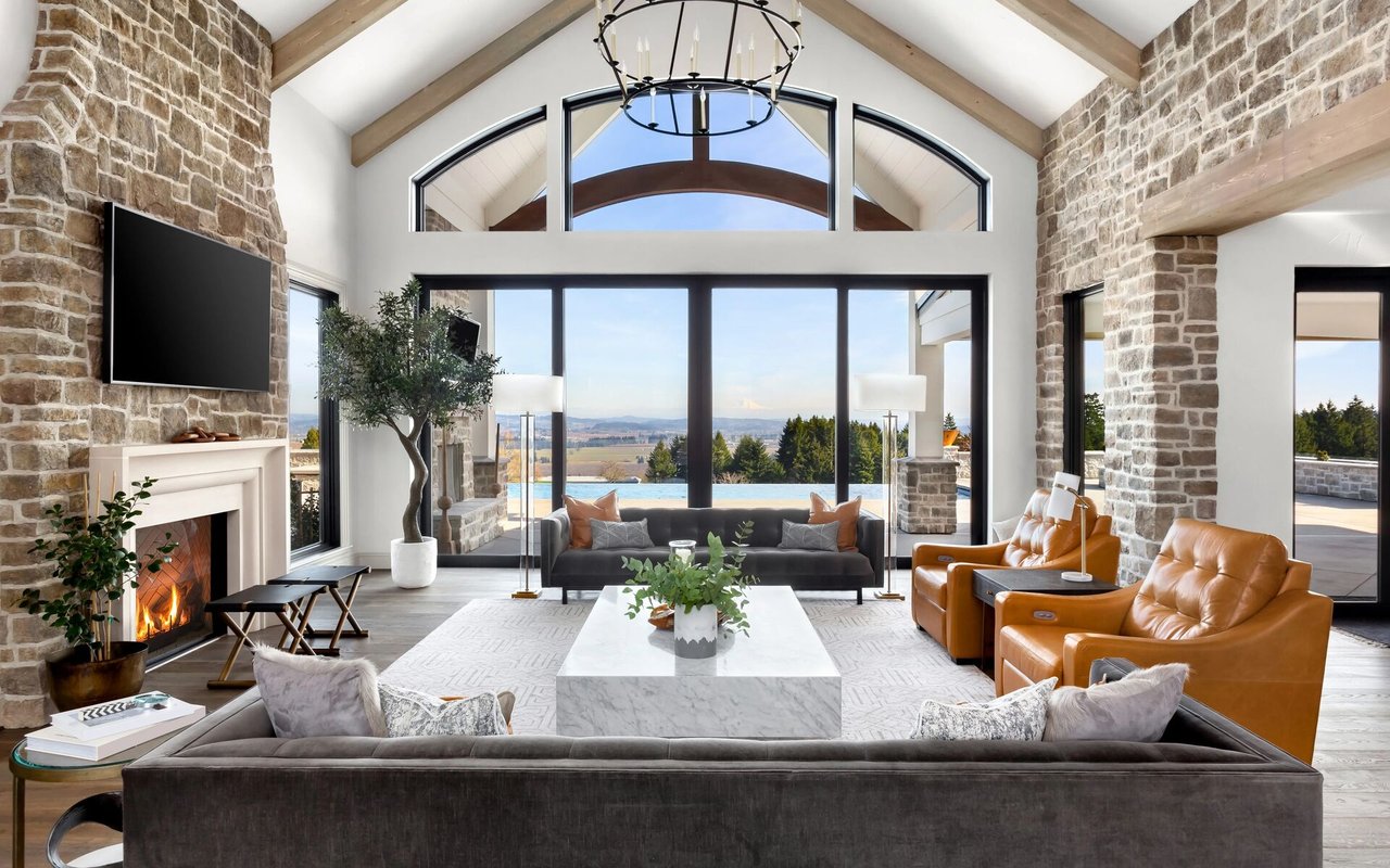 Luxury living room with vaulted wood-beam ceiling, stone fireplace, and arched windows overlooking a pool.