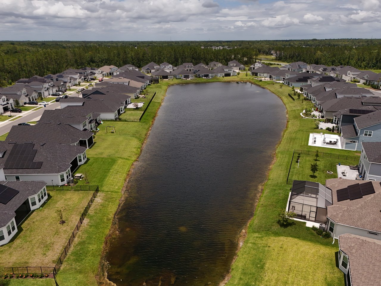 Aerial view of homes lining a central pond, with green lawns and woods beyond.