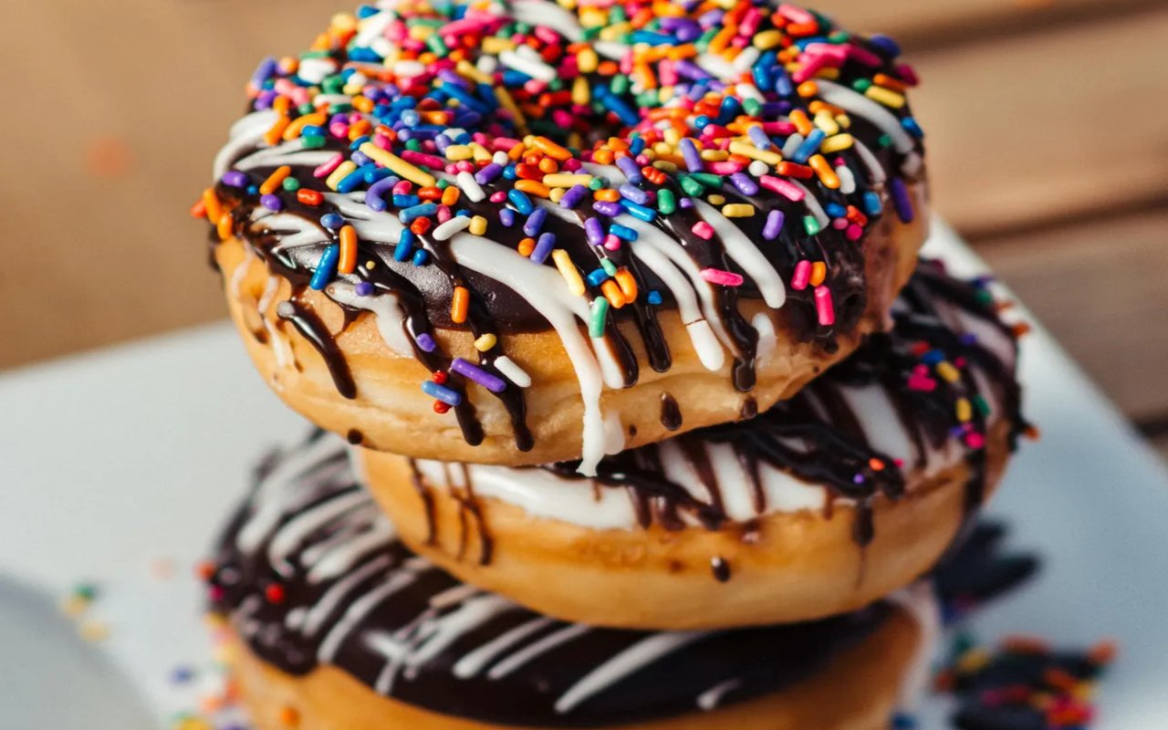 A close-up of a stack of three chocolate-frosted donuts with white icing drizzle and colorful sprinkles on top.