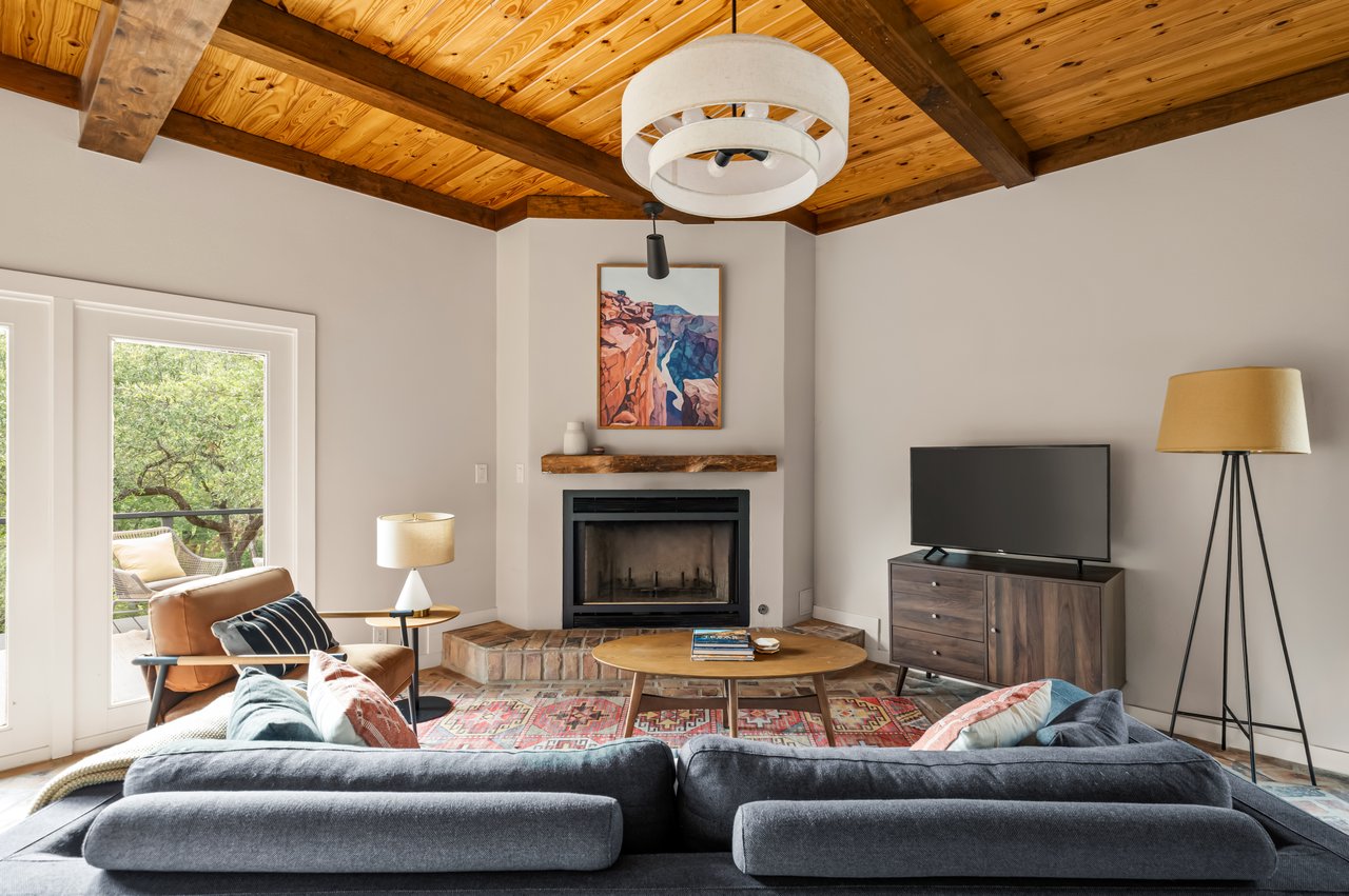 Living room with a vaulted wood ceiling, brick fireplace, and glass doors opening toward trees.