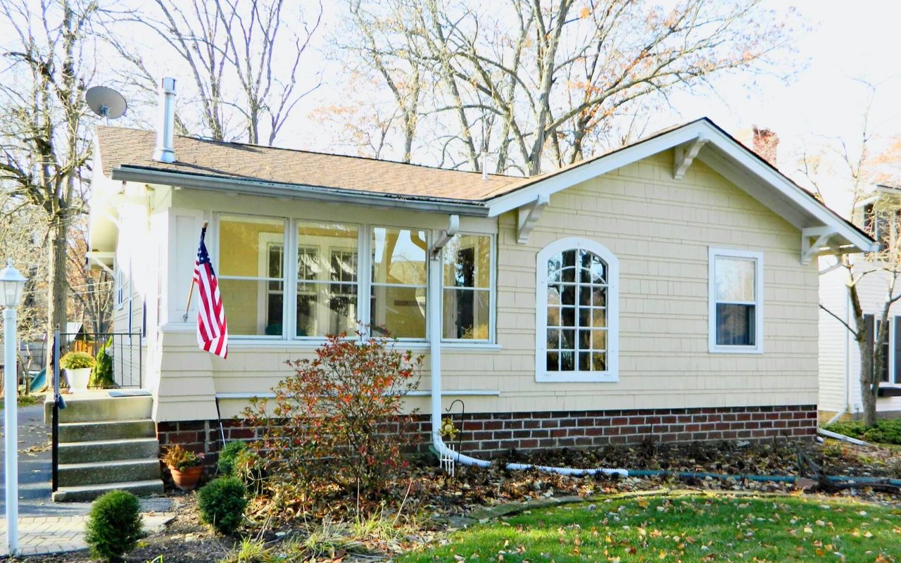 Light yellow single-story house with a covered porch, brick foundation, and American flag.