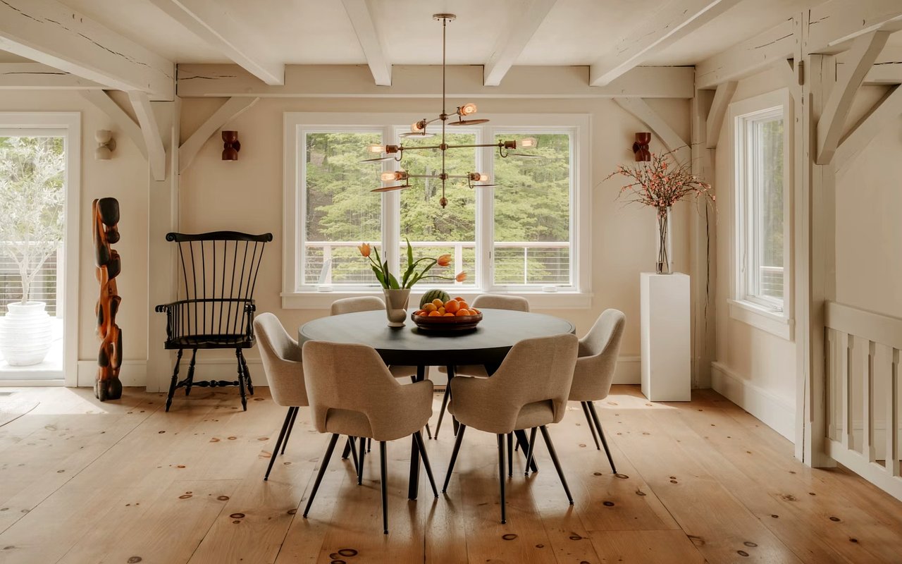 Dining room with exposed beams, a round table, and large windows overlooking trees.
