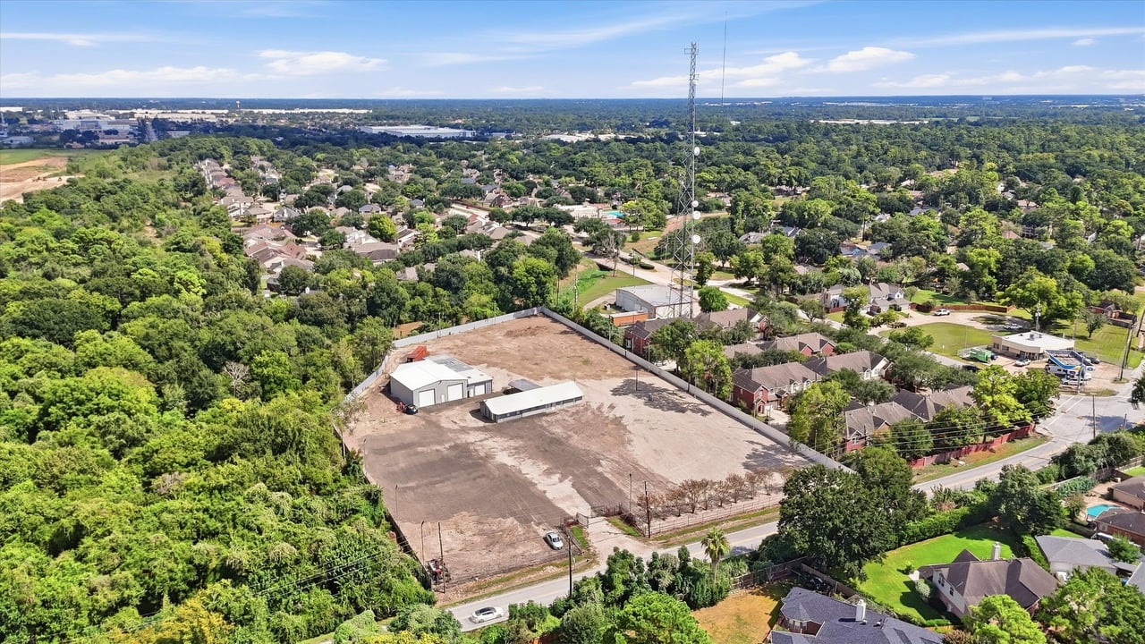 Aerial view of a cleared fenced lot with two buildings and a communications tower among nearby homes.