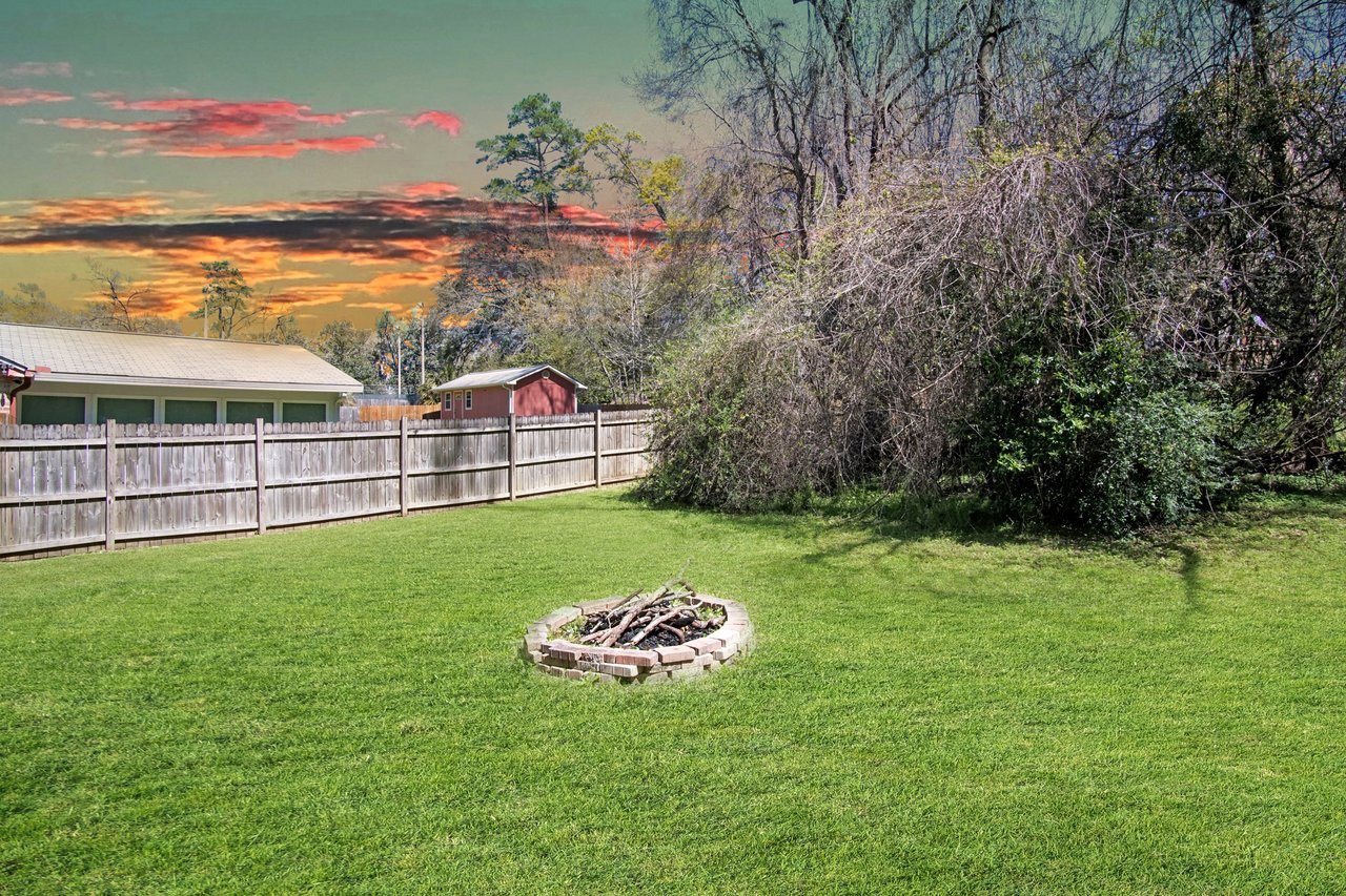 Spacious backyard with a green lawn, a circular stone fire pit, and a wooden fence. The sunset sky casts a warm glow over the scene.