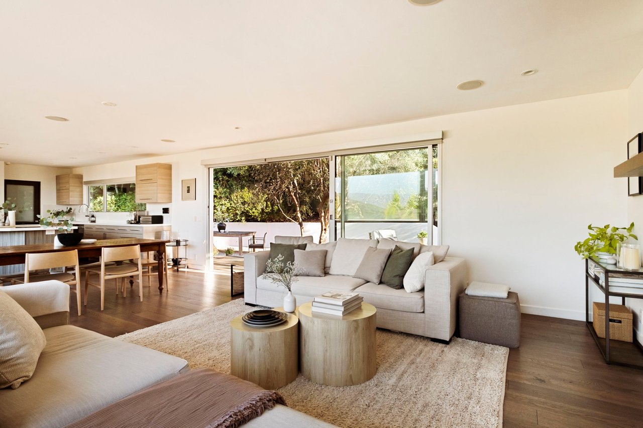Open-plan living room with neutral sofas, light wood floors, and sliding glass doors to a leafy patio.