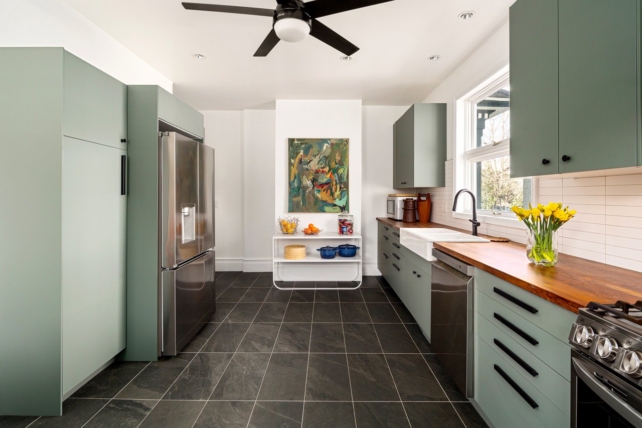 Modern kitchen featuring sage green flat-panel cabinets, butcher block countertops, and stainless steel appliances set against dark tile flooring.